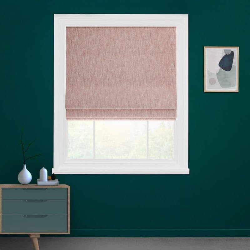 Blush-pink Roman shade inside a white window frame, partially lowered and filtering daylight. Set on a dark teal wall in a minimalist room with a dresser and framed abstract art.