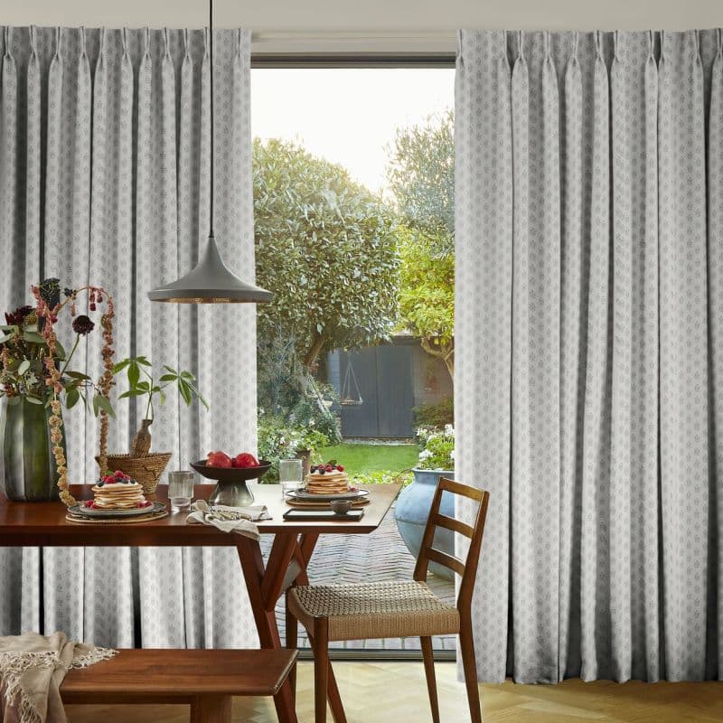Patterned pleated curtains, pale grey, pulled aside revealing a sliding glass door; filtering soft daylight into a dining nook with wooden table set for breakfast and a green garden view.