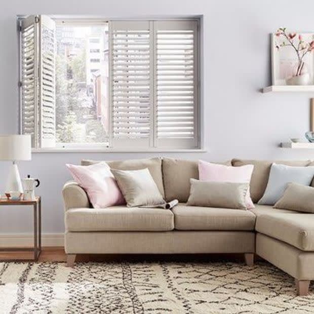 White plantation shutters, three-panel with one side folded open, slats partially closed, filtering daylight through an urban view; mounted above a beige L-shaped sectional in a softly lit living room.