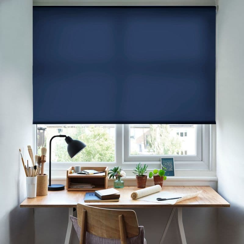 Navy roller blind, fully lowered across a double window, blocking light; covering a small wooden desk with lamp, brushes, notebooks and potted plants in a bright home workspace.
