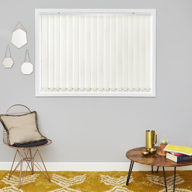 Cream vertical blinds (inside window frame, scalloped weighted bottoms) — closed, softly diffusing daylight — in a grey-walled living room with a wire chair, round wooden coffee table, and mustard patterned rug.