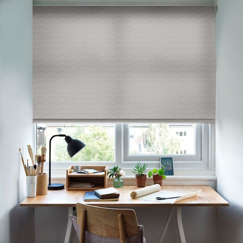 A light-gray roller blind, fully lowered across a double window, filtering soft daylight; a wooden desk beneath holds a lamp, brushes, notebooks and potted succulents in a bright workspace.