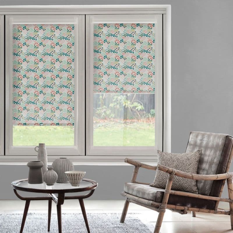 Pastel floral roller blinds inside two casement frames — left fully lowered, right partially raised — filtering daylight in a grey-walled room with rattan armchair, patterned cushion, coffee table and backyard view.