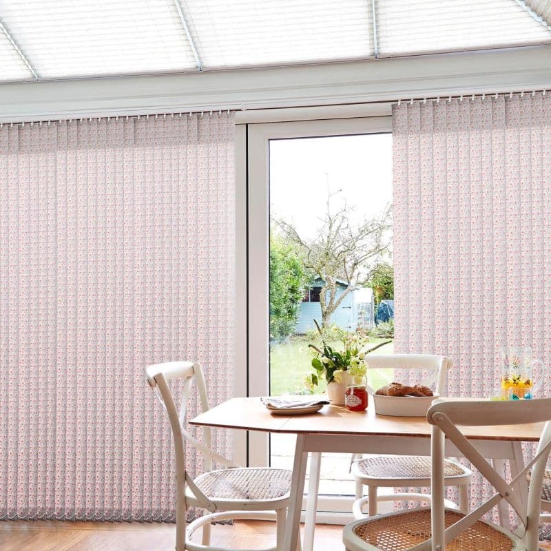 Patterned vertical fabric blinds, hanging from a ceiling track, mostly closed with a central opening, filtering soft daylight over a small dining table and chairs facing a sunlit backyard.