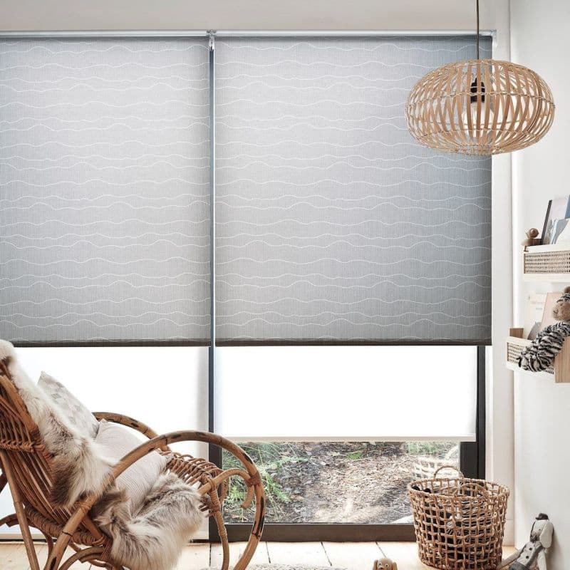 Light-gray patterned roller blinds (two adjacent panels with subtle white wavy lines), fully lowered and diffusing daylight over a sliding window; cozy nook with rattan chair, wicker pendant, basket, floor.