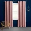 Textured pink grommet-top floor-length curtains hang slightly parted on a metal rod, framing daylight from a single-pane window in a navy-walled room with a gold wire chair and circular sconce.