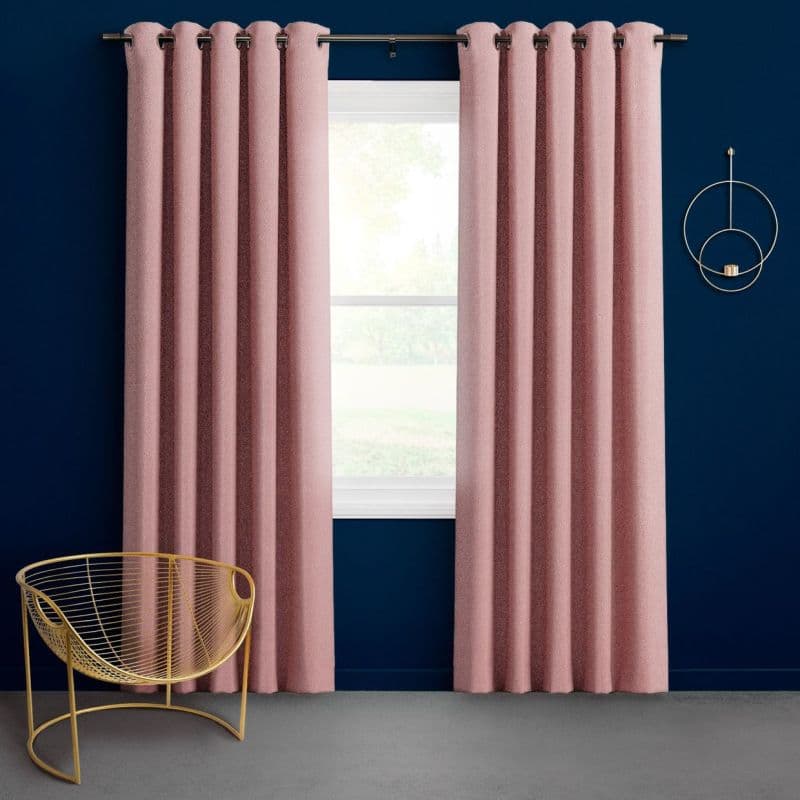 Textured pink grommet-top floor-length curtains hang slightly parted on a metal rod, framing daylight from a single-pane window in a navy-walled room with a gold wire chair and circular sconce.