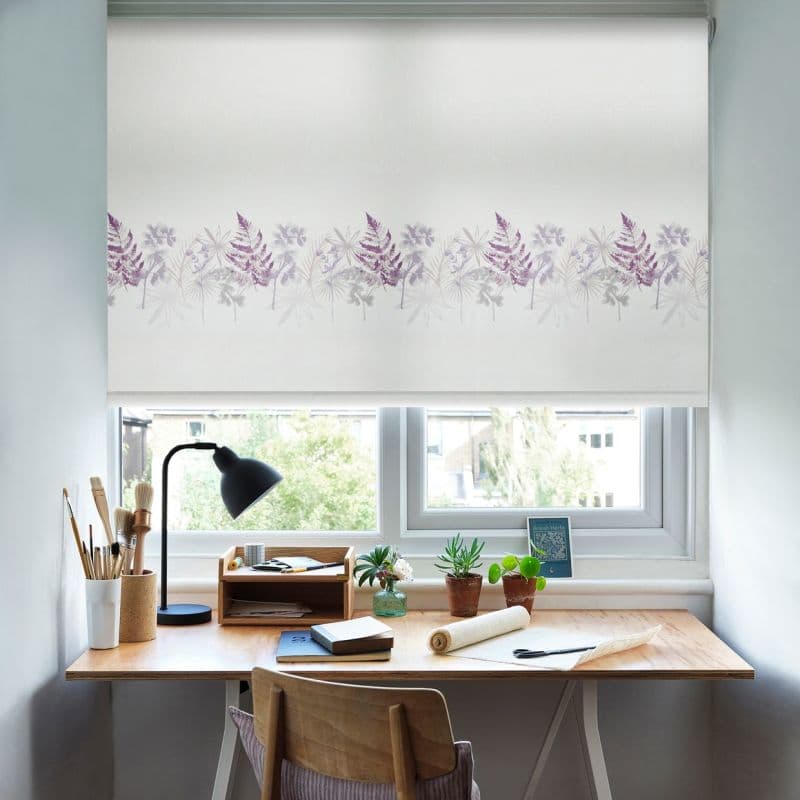 White roller blind with a horizontal purple fern-and-flower border, lowered and filtering daylight; sits over a double window above a wooden home desk with lamp, plants, notebooks, and city-tree view.