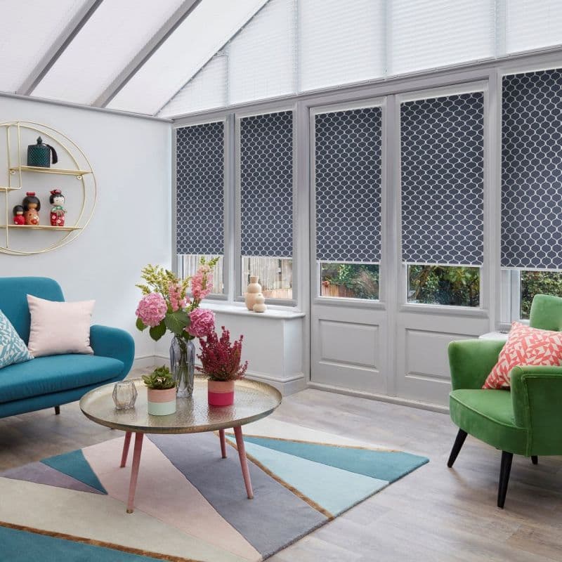 Patterned navy-and-white roller blinds covering tall glazed doors, lowered to mid-height and filtering light; set in a bright conservatory-style sitting room with teal sofa, green armchair and round coffee table.