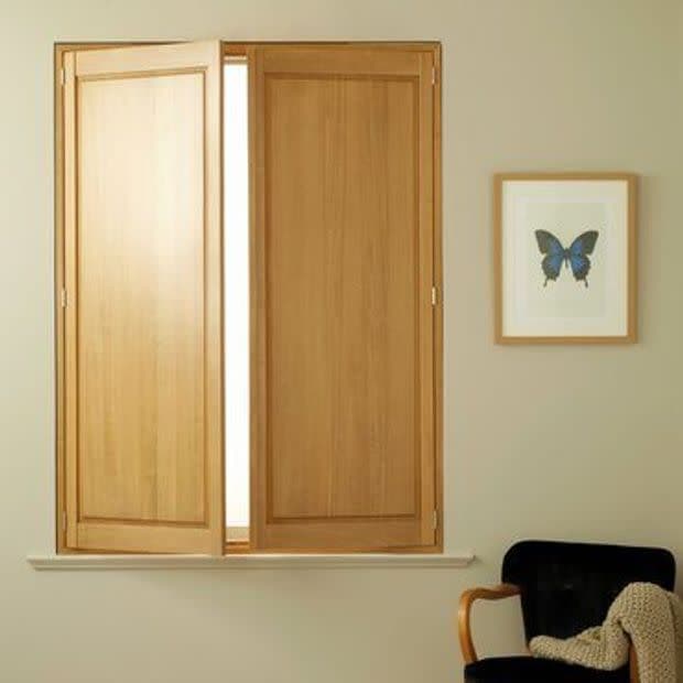 Interior oak shutters—two hinged panels—cover a rectangular window; one panel is slightly ajar, admitting bright light into a neutral-toned room with a framed blue butterfly print and a black armchair.