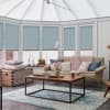 Light-blue pleated cellular blinds, lowered over tall conservatory windows with uncovered bottoms; filtering soft daylight; in a bright, cozy living area with beige sofa, wooden coffee table and rug.