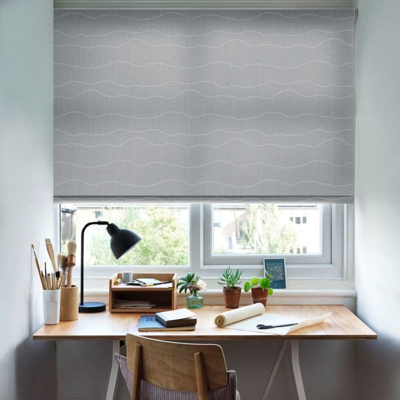 Grey patterned roller blind, lowered over the window with wavy white lines across the fabric; filtering soft daylight into a compact home workspace — wooden desk with lamp, plants, and art supplies.