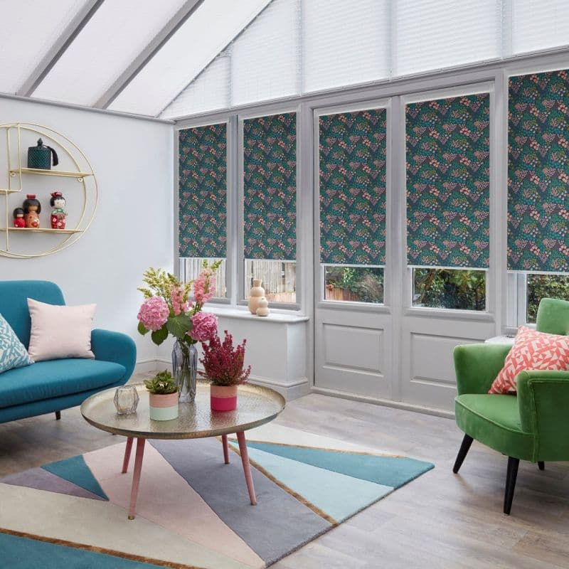Floral patterned roller blinds covering tall conservatory windows and doors, lowered to mid-height and filtering daylight; contemporary sunroom with pastel sofas, geometric rug, coffee table and potted flowers.