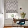 Chevron-patterned Roman blind, white with black and tan dotted chevrons, lowered over the window, filtering daylight; small bright home office with wooden desk, teal chair, vase of flowers, garden view.