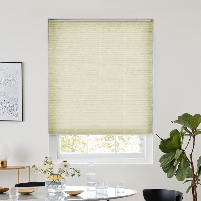Light-yellow pleated cellular shade, fully lowered over a sash window, filtering daylight; set in a bright minimalist dining area with round table and vase, framed art left, fiddle‑leaf fig right.