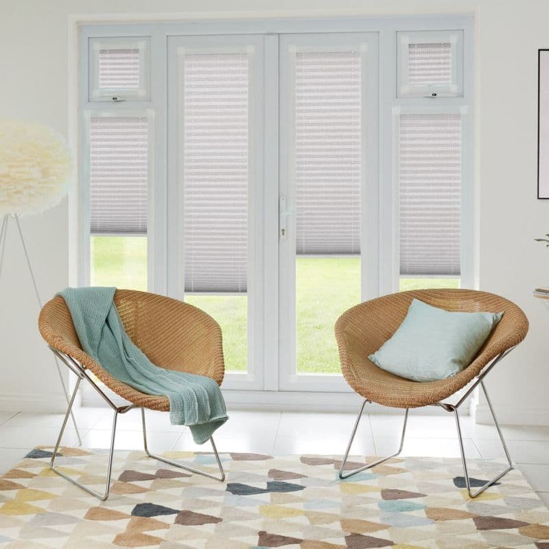 Pleated cellular blinds on French doors and sidelights, light-gray and lowered mid-height, filtering daylight; bright sitting area with two woven rattan chairs, pastel cushions, patterned rug and lawn view.