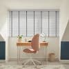 White horizontal slatted blinds with grey vertical tapes cover a wide dormer window, closed and filtering soft daylight; small home study with wooden desk, pink swivel chair and lamp.