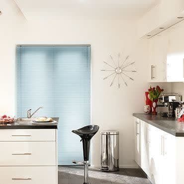 Light-blue horizontal Venetian blinds covering a tall window, fully closed and softly diffusing daylight; bright modern kitchen with white cabinets, countertop appliances, bar stool, trash bin, and wall clock.