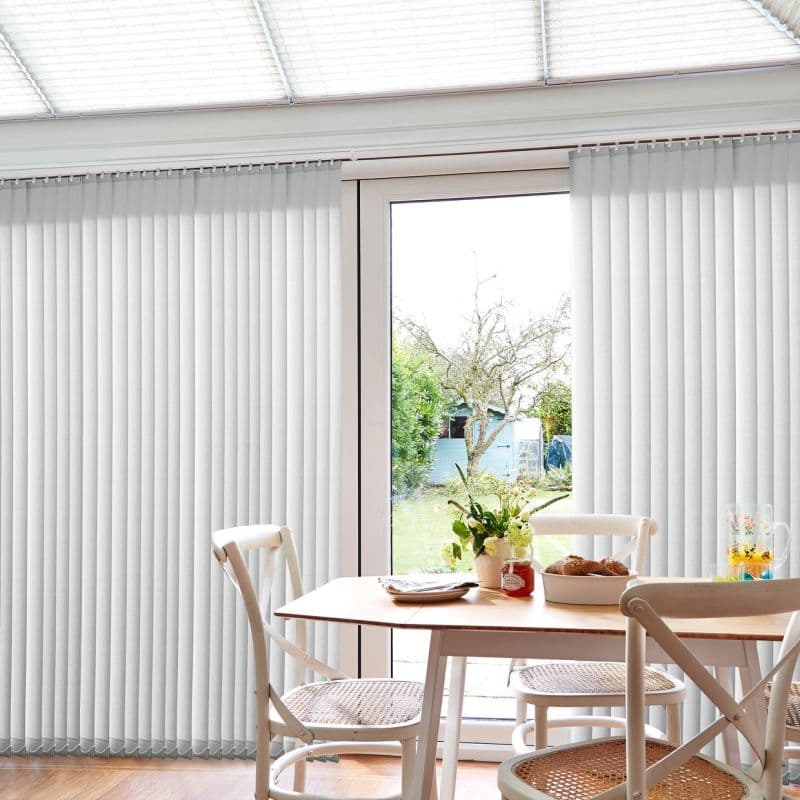 White vertical blinds, floor-to-ceiling and evenly spaced, parted centrally to expose a glass patio door, filtering soft daylight into a dining nook with wooden table, chairs, and garden view.