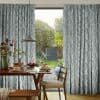 Floor-to-ceiling curtains with blue-grey vertical brushstroke pattern, parted to reveal a sunlit garden and sliding glass door. Inside, a wooden dining table set for breakfast beneath a pendant lamp.
