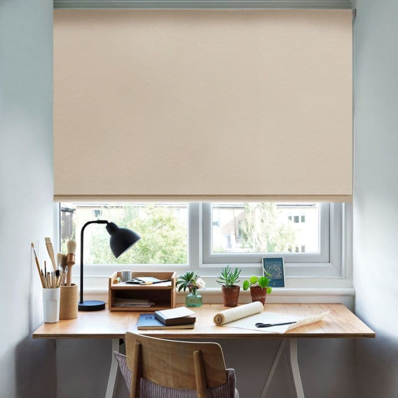 Beige fabric roller blind, lowered almost to the sill with a smooth texture; filtering soft daylight and obscuring the view over a wooden desk with lamp, brushes, notebooks, and potted plants.