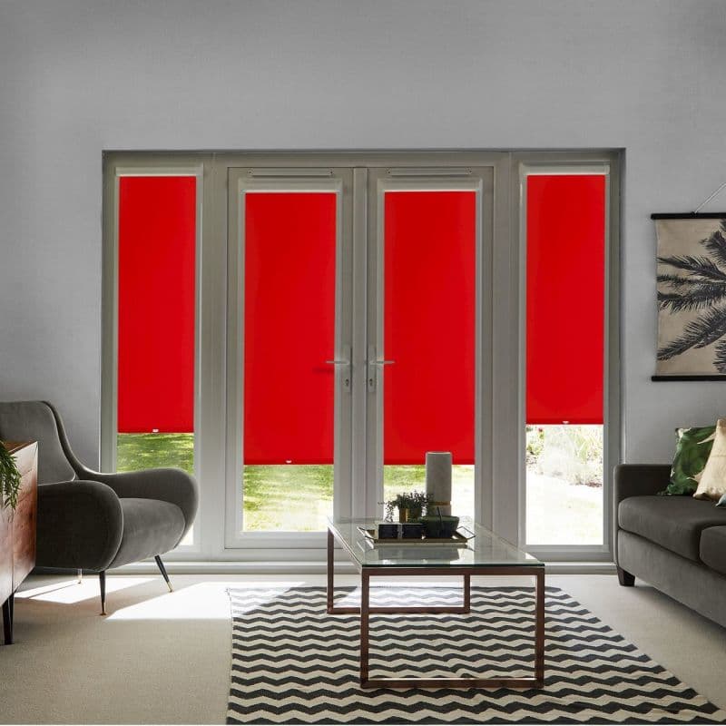 Red roller blinds fill four-panel French doors and sidelights, fully lowered and opaque with slim bottom gaps revealing lawn; contemporary living room with gray sofa and glass coffee table.