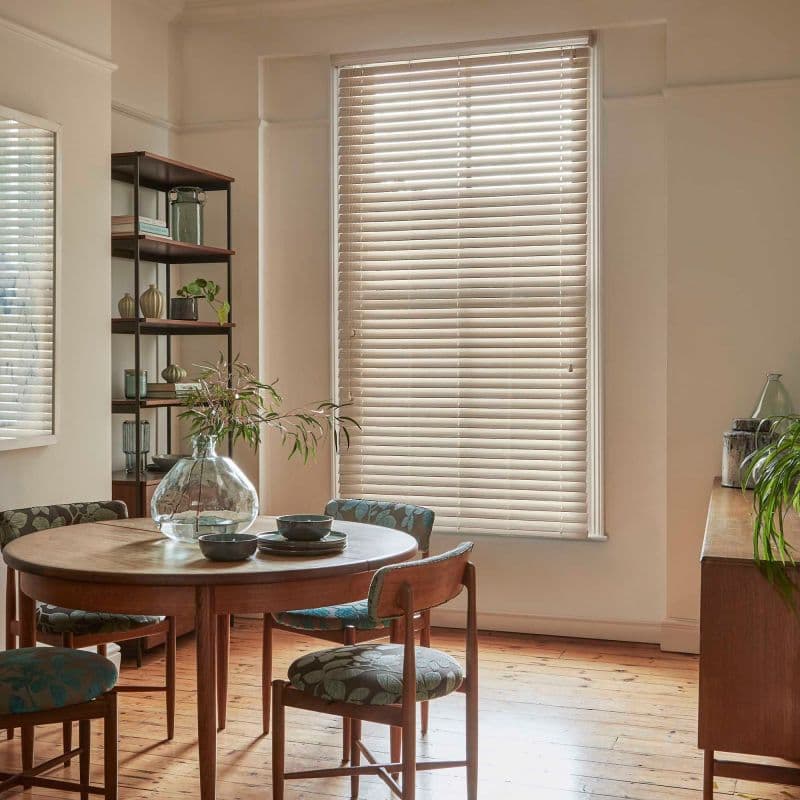 Beige horizontal blinds covering a tall window, closed and gently filtering daylight; in a sunlit dining room with a round wooden table, patterned chairs, open shelving, plants, and hardwood floor.