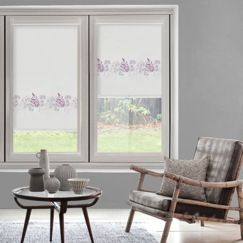 Two white roller blinds with a lavender fern border—one fully lowered, one partly raised—filtering daylight in a gray living room with a rattan armchair, side table, and a grassy garden view.