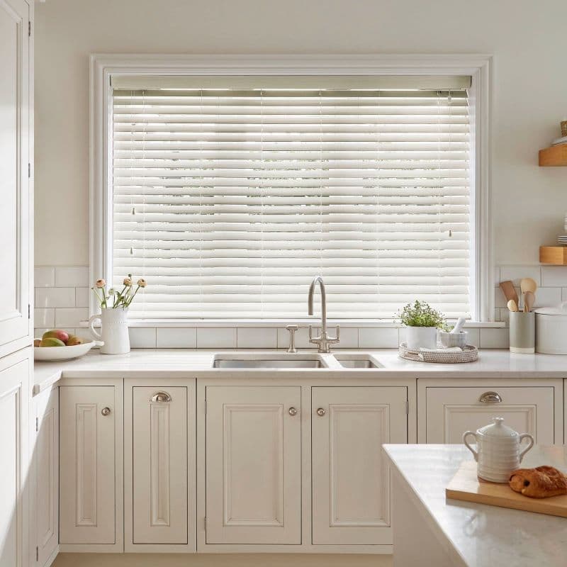 White horizontal Venetian blinds covering a wide window; slats closed, softly filtering daylight; positioned above a double stainless sink on a marble countertop in a bright, neutral kitchen with cream cabinetry and plants.