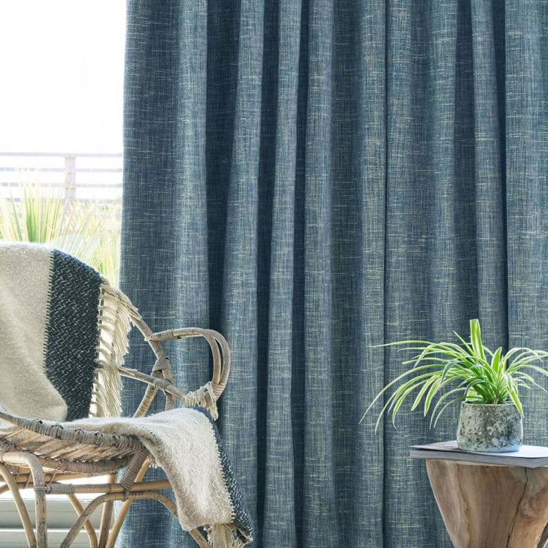 Textured teal curtains, floor-to-ceiling and mostly closed with a subtle crosshatch weave, filtering daylight; a wicker chair draped with a throw and a wooden side table holding a potted plant.