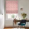 Coral-and-white patterned Roman blind, lowered and filtering daylight over the window; a wooden desk beneath holds a round mirror, vase of flowers, scarf and objects in a bright neutral home office.
