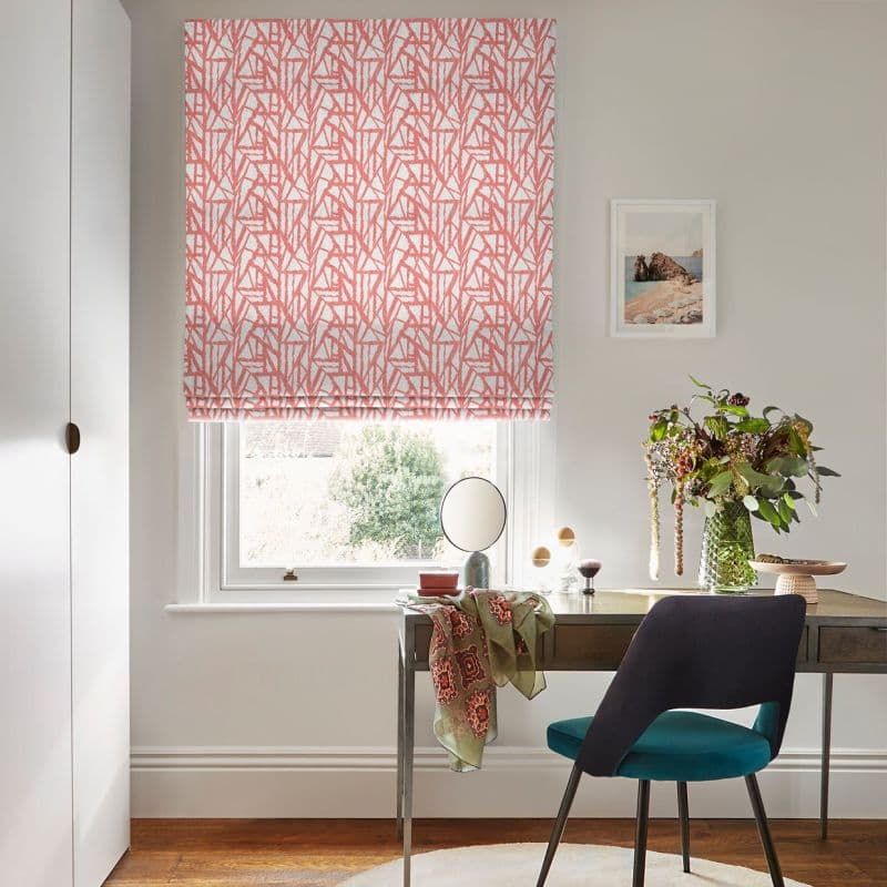 Coral-and-white patterned Roman blind, lowered and filtering daylight over the window; a wooden desk beneath holds a round mirror, vase of flowers, scarf and objects in a bright neutral home office.