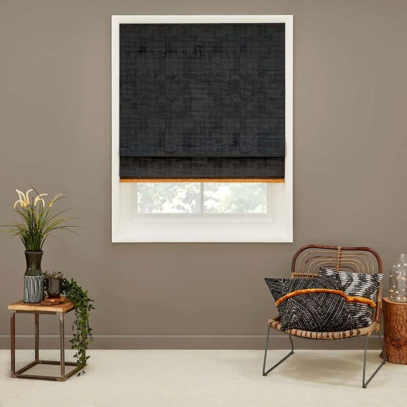 Dark charcoal Roman blind lowered over window, orange trim at hem, filtering daylight. In a neutral-toned living corner with a rattan chair, patterned cushions, small side table and potted plants.