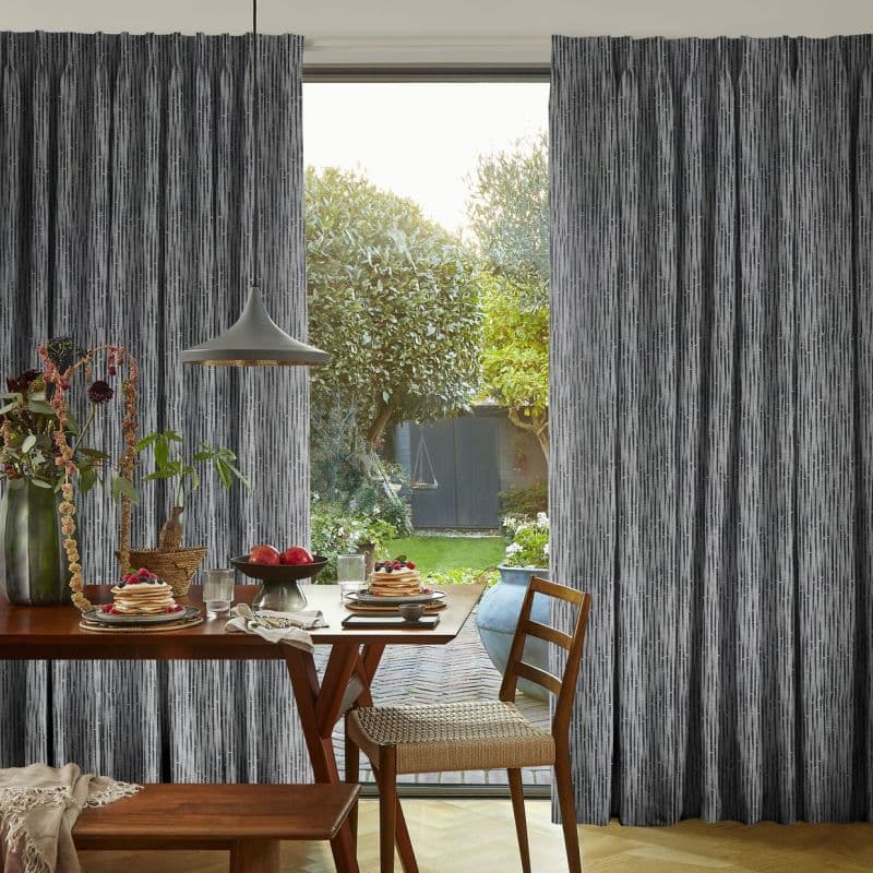 Full-length grey textured curtains with vertical striations, parted at center; framing and filtering sunlight onto a wooden dining table with pancakes and fruit, lamp and chairs, opening onto a garden.