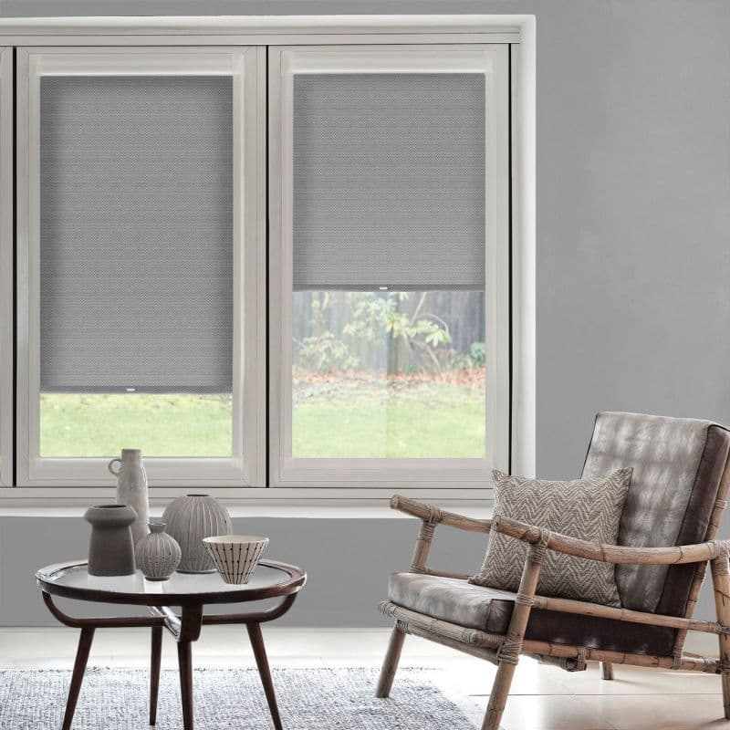 Gray textured roller blinds, partially lowered and filtering light on tall windows; living room with rattan leather armchair, round coffee table with ceramic vases, and a garden visible outside.