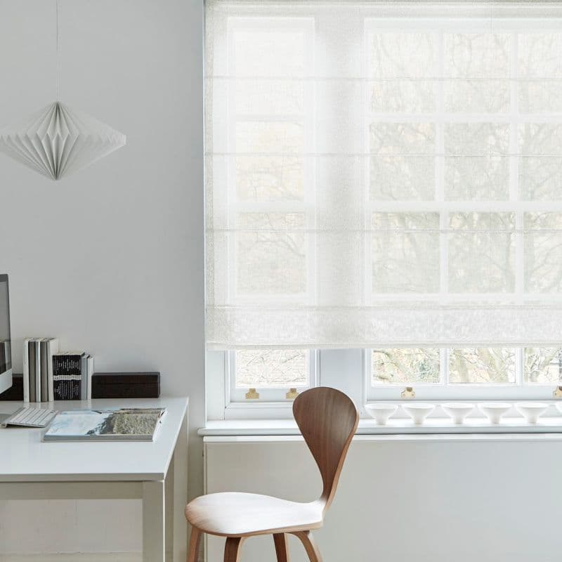 White sheer Roman shade, lowered across a multi-pane window with soft horizontal folds, filtering diffuse daylight; minimalist home office with white desk, wooden chair, pendant lamp, and bare-tree view.