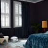 White plantation shutters on two tall windows, paired with dark floor-length curtains drawn to one side; shutters closed, filtering sunlight into a dim, moody bedroom with velvet bedspread, lamp, plant.