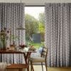 Patterned pleated curtains, floor-length with dotted chevron print, pulled partly aside to reveal a glass sliding door; dining table set for breakfast under a pendant lamp, sunlit garden beyond.
