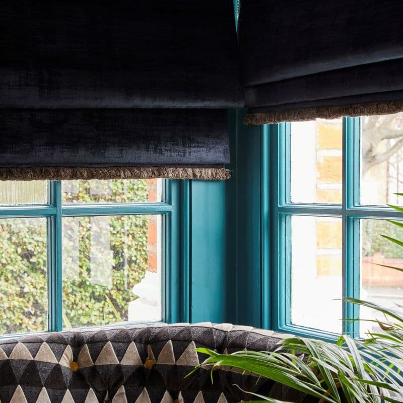 Dark velvet Roman blinds with fringed trim, partially lowered and dimming daylight, over turquoise-painted mullioned corner windows in a room with patterned seating, a plant; brick and garden visible outside.