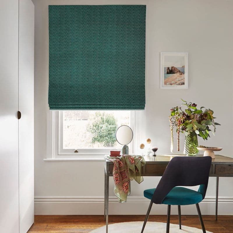 Teal patterned Roman blind lowered over a sash window, blocking light and filtering soft daylight; small desk/vanity beneath with mirror, vase of flowers, chair in a bright, neutral home office.