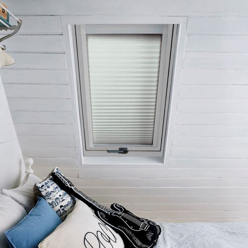 Pleated white blind in a recessed skylight, fully lowered and closed, diffusing soft daylight into a white-painted attic bedroom above a bed with blue patterned pillows and a guitar-shaped cushion.