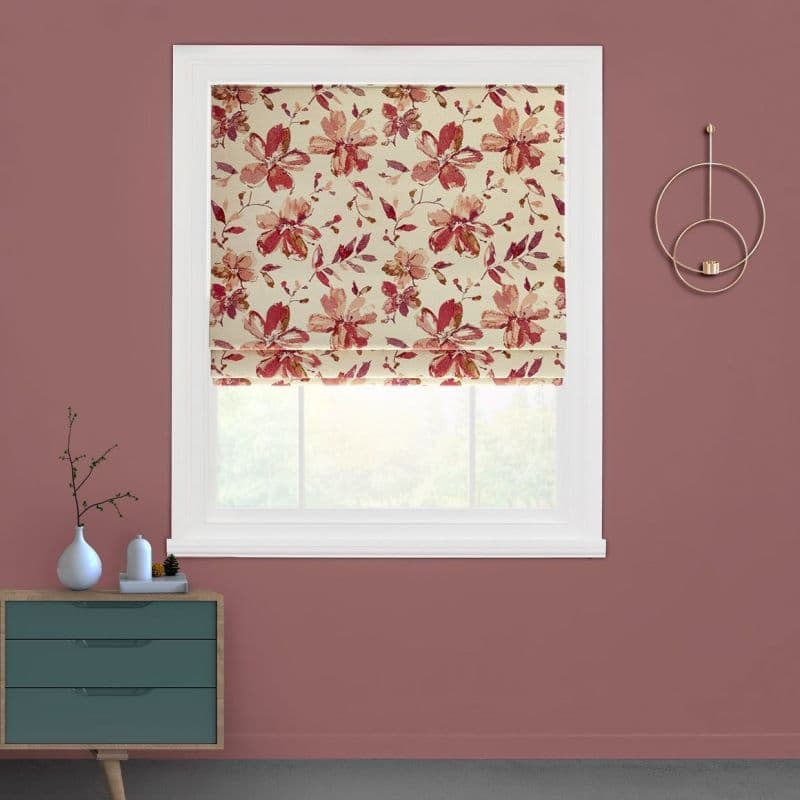 Floral Roman blind (cream with pink-red blossoms), partially lowered with visible folds, filtering soft daylight through a white-framed window in a mauve room with teal dresser and minimal decor.