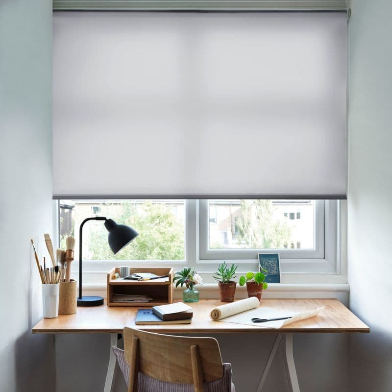White roller blind, pulled down to cover the upper three-quarters of a double window, smooth translucent fabric softly filtering daylight; above a wooden desk workspace with lamp, potted plants, notebooks and chair.