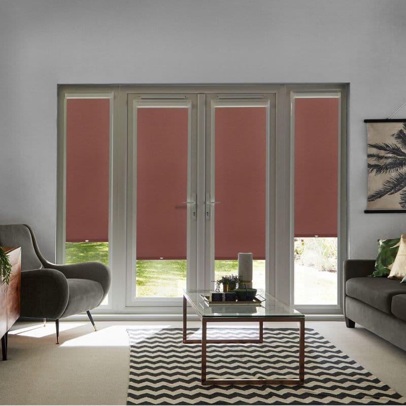 Solid muted-red roller blinds, lowered across full-height French doors and sidelights with small bottom gaps, filtering daylight; living room with grey sofas, glass coffee table, chevron rug, sunlit lawn beyond.