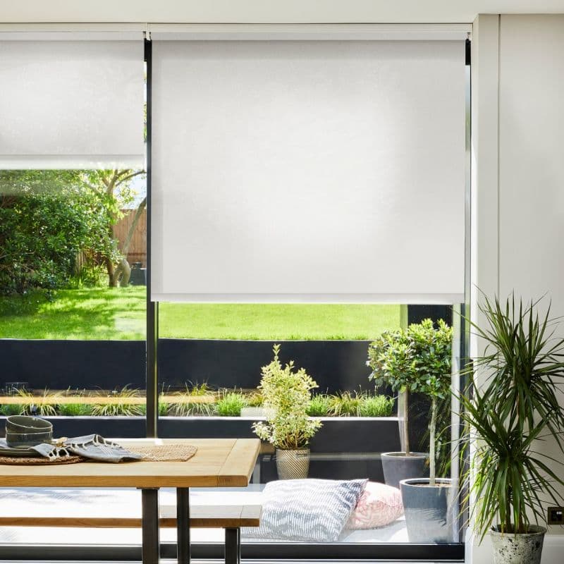 White roller blind, lowered mid-window with slight translucence, filtering daylight; overlooks sunlit backyard with lawn and potted plants, dining table and bench in the foreground.