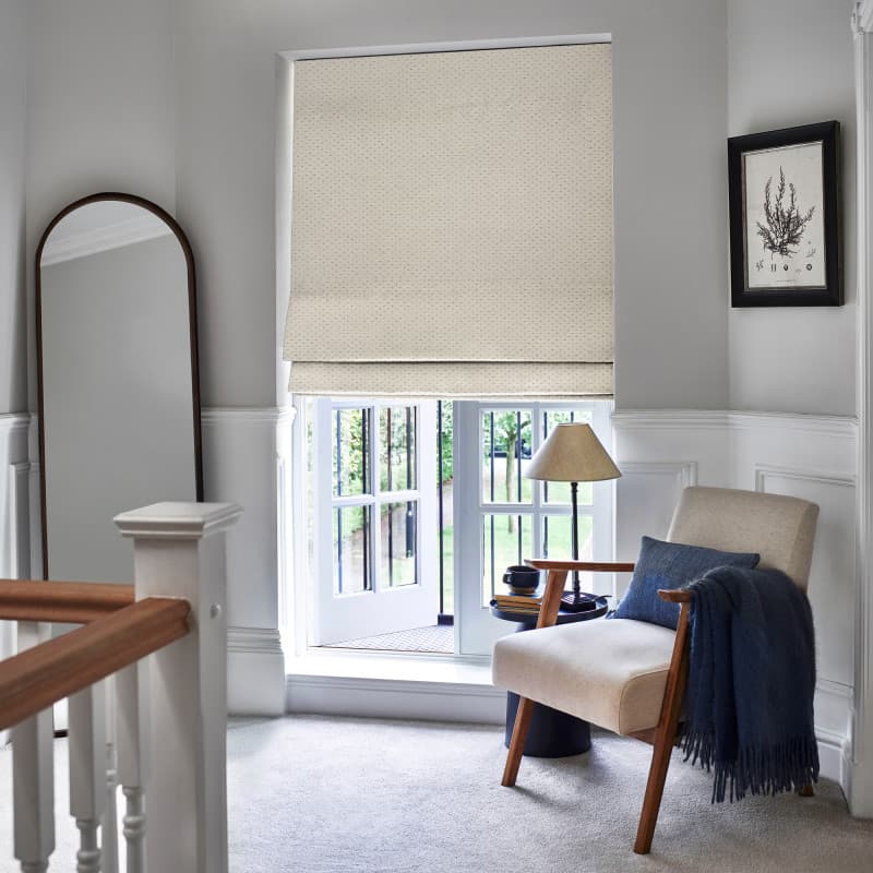 Beige dotted Roman shade lowered over window, softly folded at bottom, filtering daylight into a bright upstairs landing nook with upholstered armchair, lamp, full-length mirror and garden view.