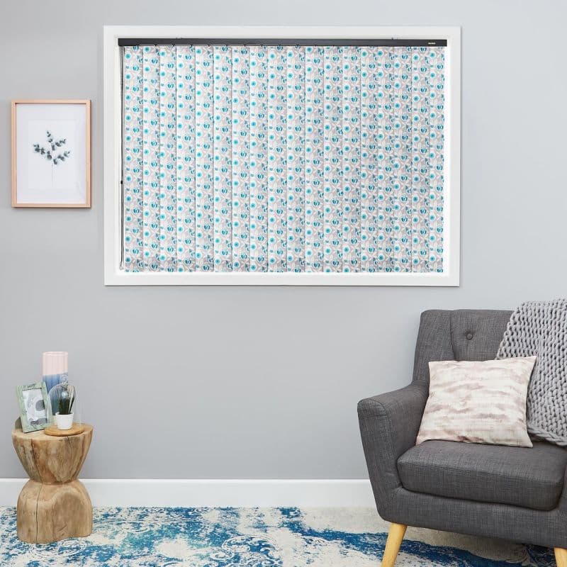 Patterned fabric vertical blinds (blue floral), fully closed and filtering light inside a white-framed window, in a grey living room with armchair, side table, print and blue rug.
