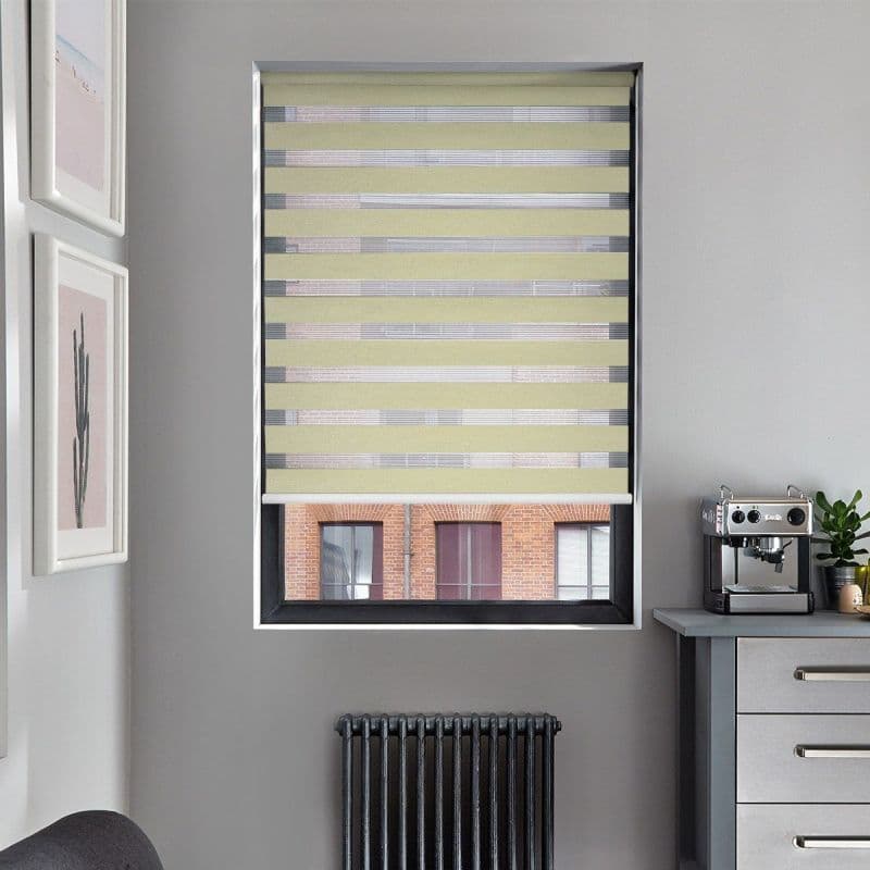 Zebra roller blind (alternating pale yellow opaque and sheer horizontal bands) lowered, filtering daylight through stripes; grey-walled kitchenette with radiator beneath, coffee machine on counter, brick building visible.