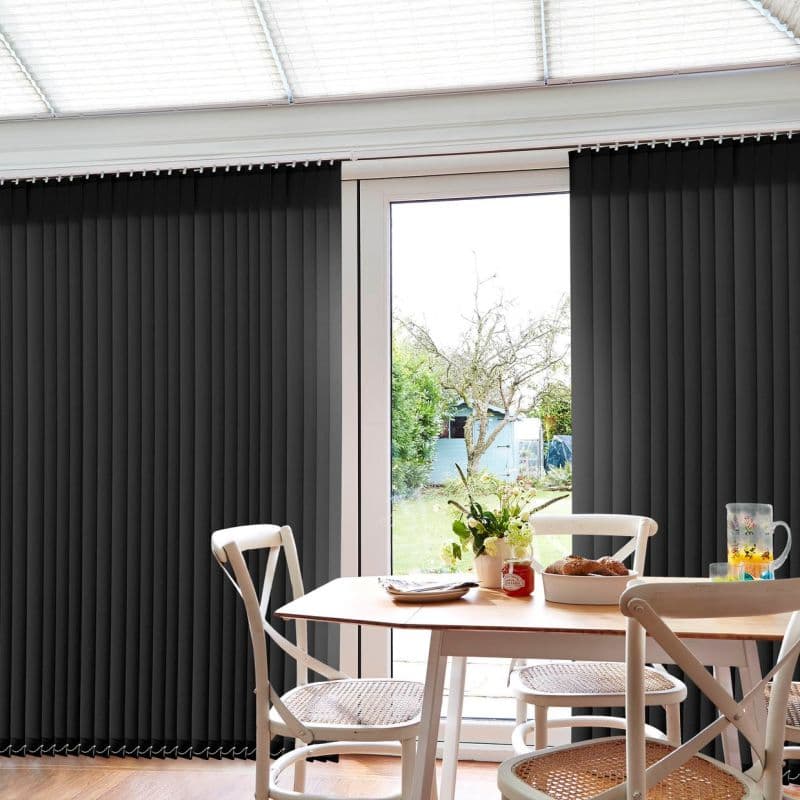 Dark grey vertical blinds spanning sliding glass doors, panels drawn to sides with central opening; filtering daylight into a small dining nook with wooden table, chairs and a garden view.