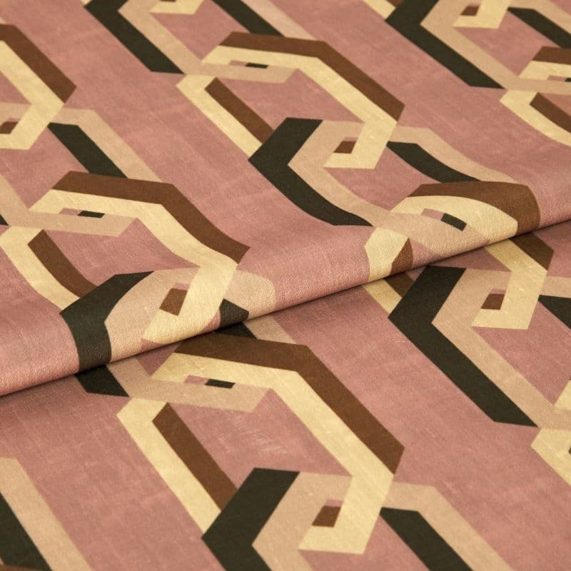 Geometric-patterned fabric with interlocking beige, brown and black hexagonal chains on a dusty rose background, folded across the center, lying on a flat surface under soft, neutral indoor lighting.